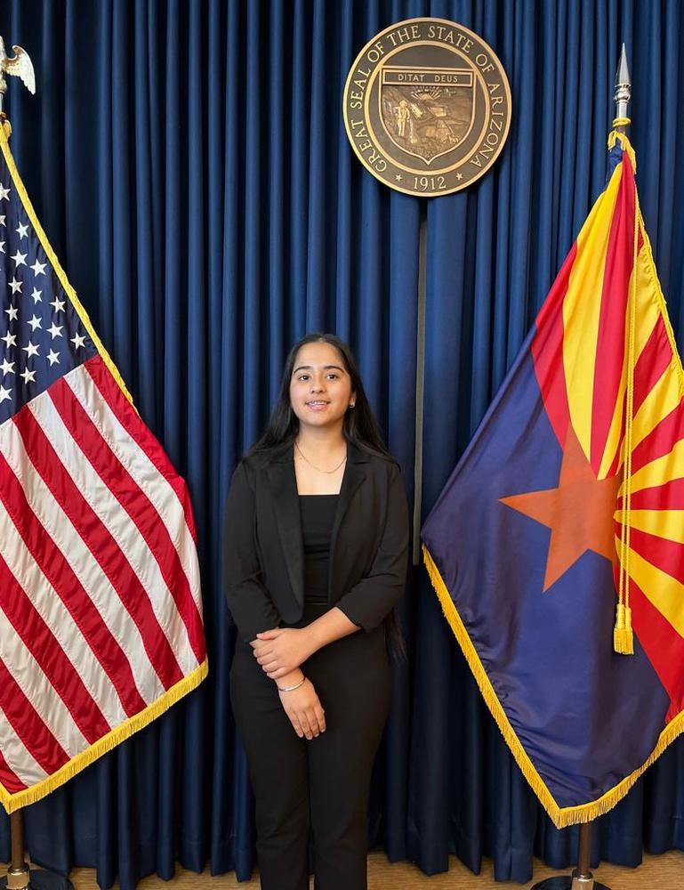 Jaisveen Kaur flanked by an American flag and an Arizona flag, standing beneath the official seal of Arizona